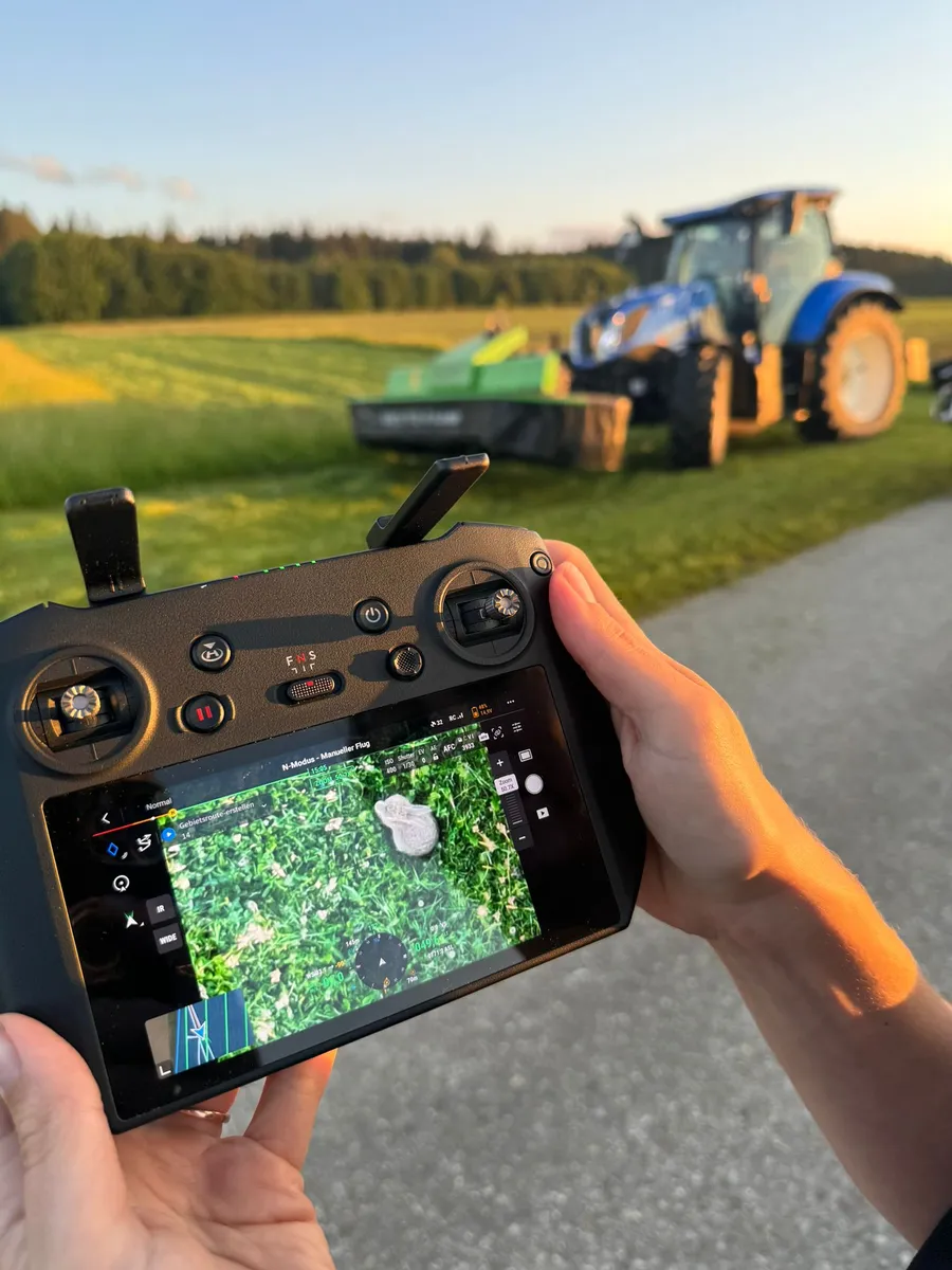 Hands holding a drone remote, its screen showing the live camera view of a fawn in a meadow; behind it, a tractor with a mower waits at the edge of the field.